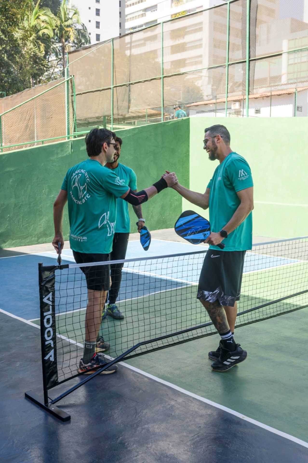 Três alunos de camiseta verde em cumprimento pós jogo de pickleball, raquete azul no centro.