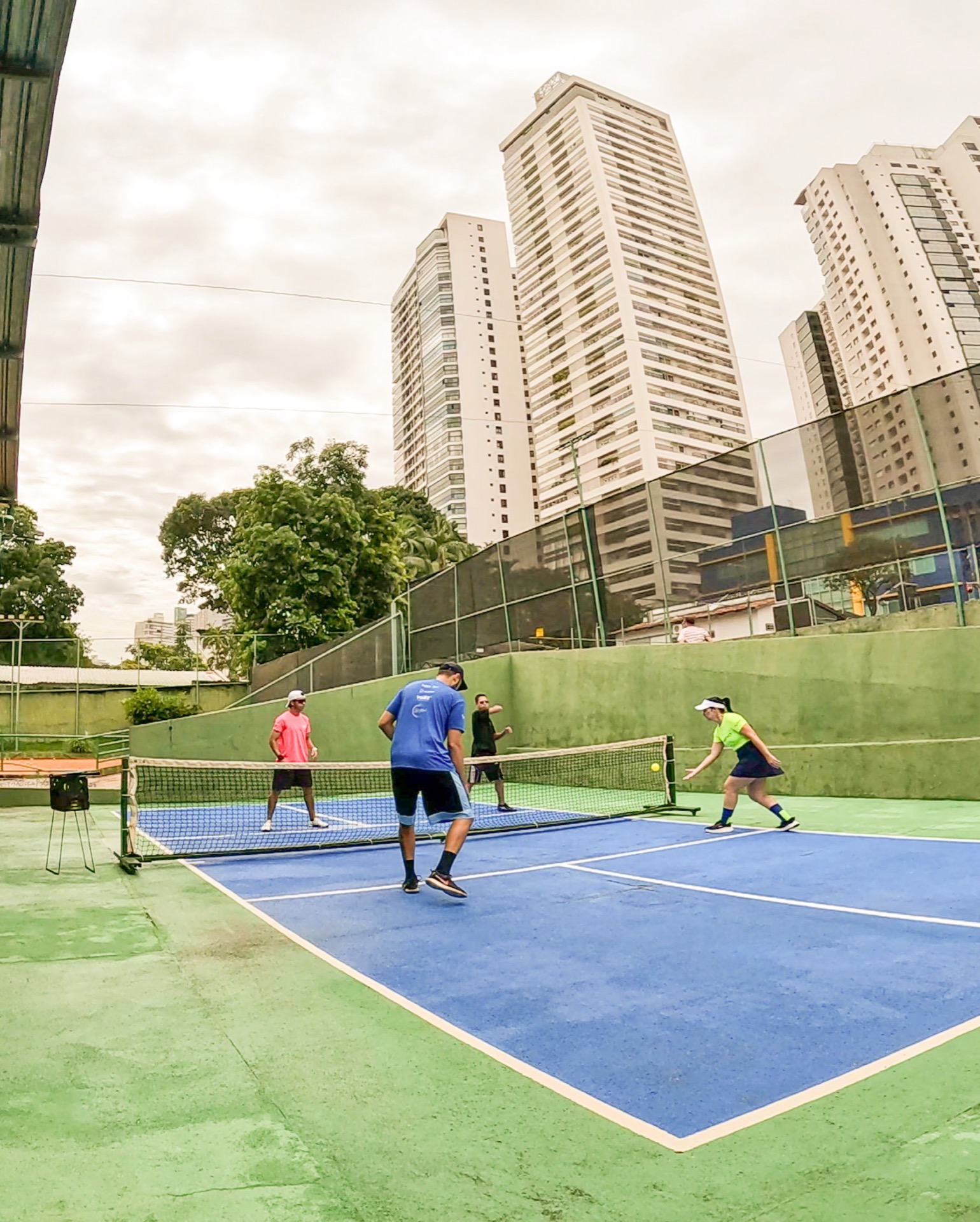 Jogo de pickleball em curso nas quadras da BTC, quatro alunos em ação, prédios de Goiânia ao fundo.