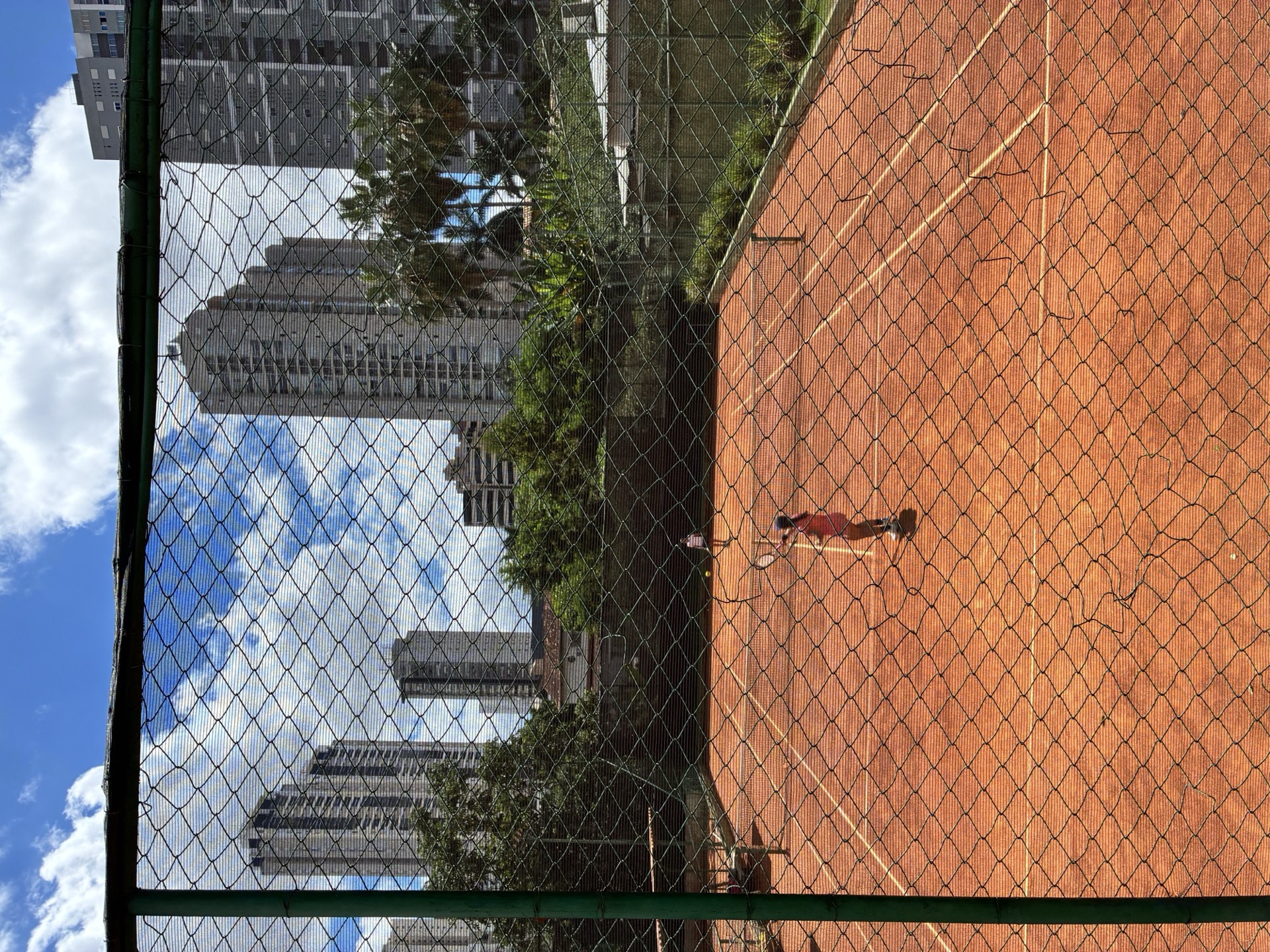 Vista da quadra de saibro da BTC pelo alambrado, prédios de Goiânia ao fundo, aluna pequena treinando no centro.