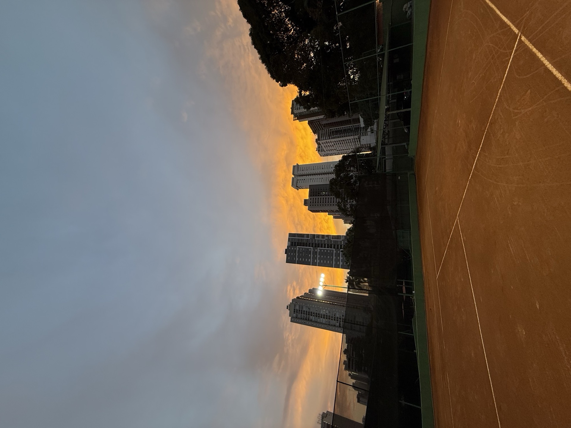 Pôr do sol dramático sobre a quadra de saibro da BTC, prédios de Goiânia em silhueta.
