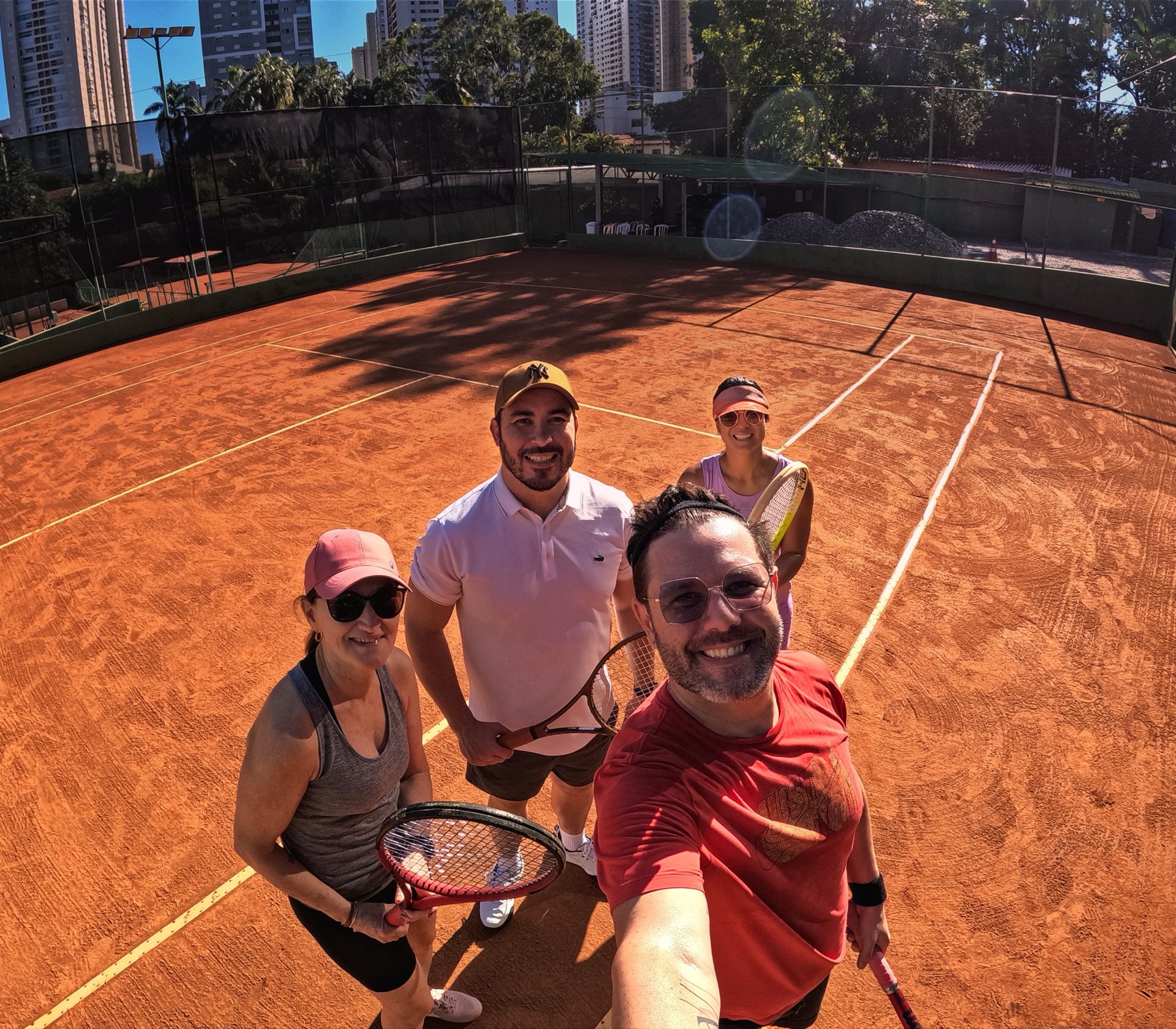 Selfie de quatro pessoas em pé na quadra de saibro da BTC, prédios de Goiânia ao fundo.